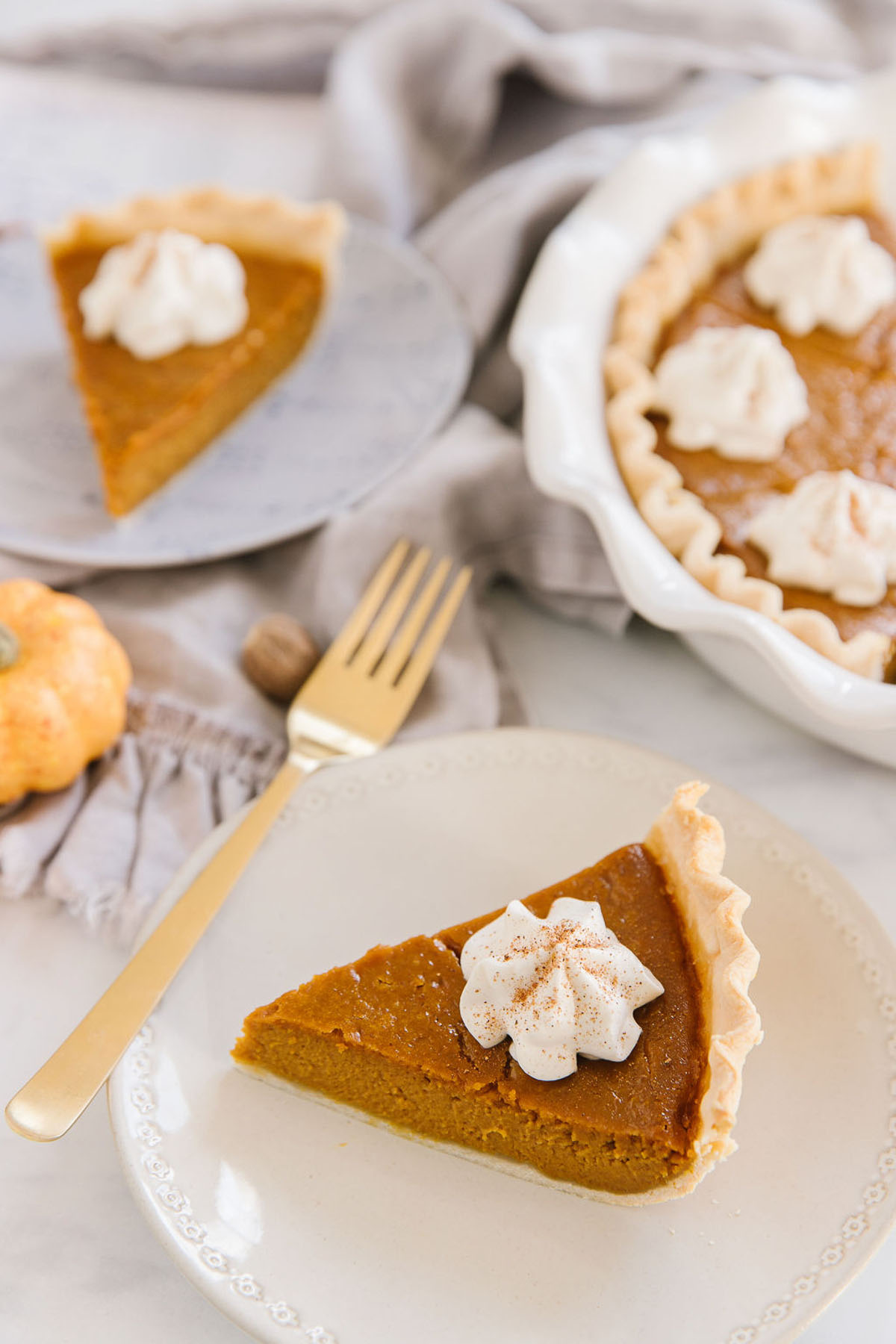 A white plate with a pumpkin pie wedge with a dollop of whipped cream on it with a gold fork on it. Behind is another pie piece on a plate and the rest of the pie in a white pan.