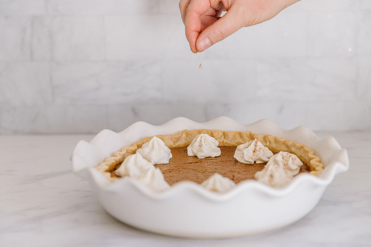 A white pan holding a fresh pumpkin pie with whipped cream dollops on it with a hand sprinkling cinnamon on it.