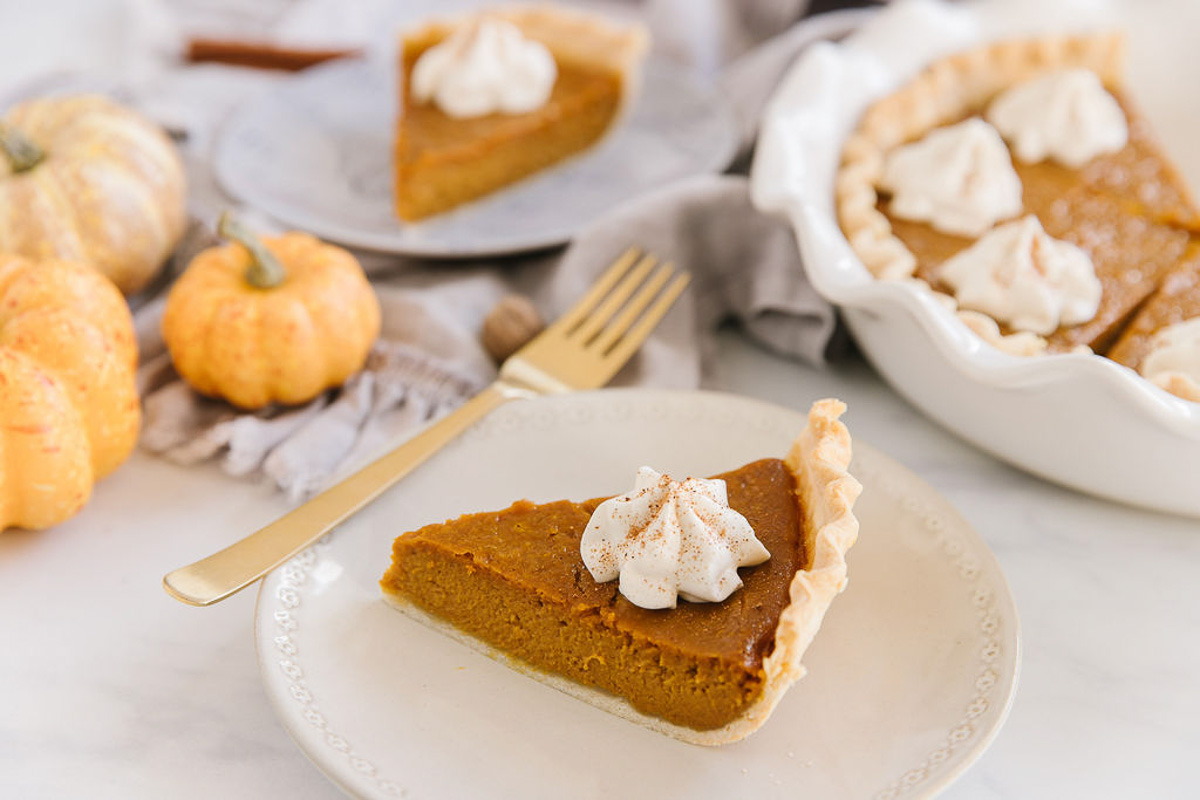 A white plate with a piece of fresh pumpkin pie with whipped cream on it with a gold fork. Behind are mini pumpkins, a blue plate holding another pumpkin pie slice and a white pan of other pumpkin pie slices.