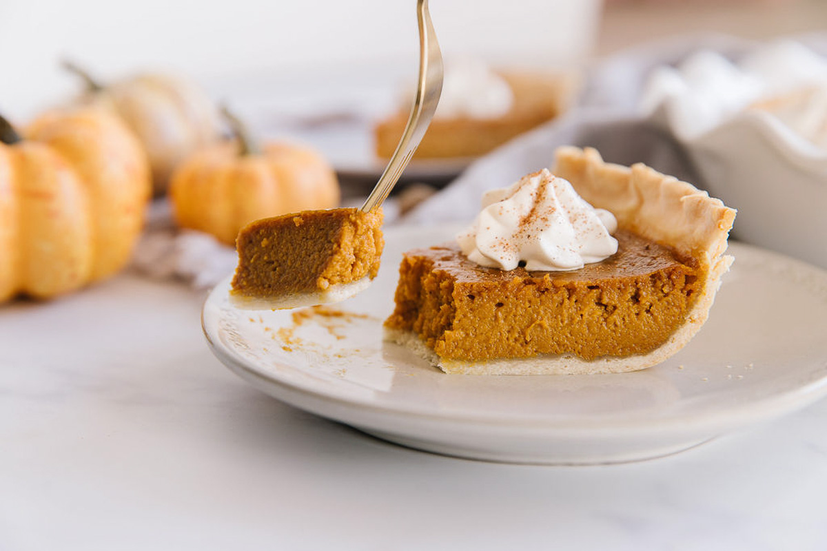 A gold fork is taking a piece of pumpkin pie from the wedge on a white plate with mini pumpkins behind it.