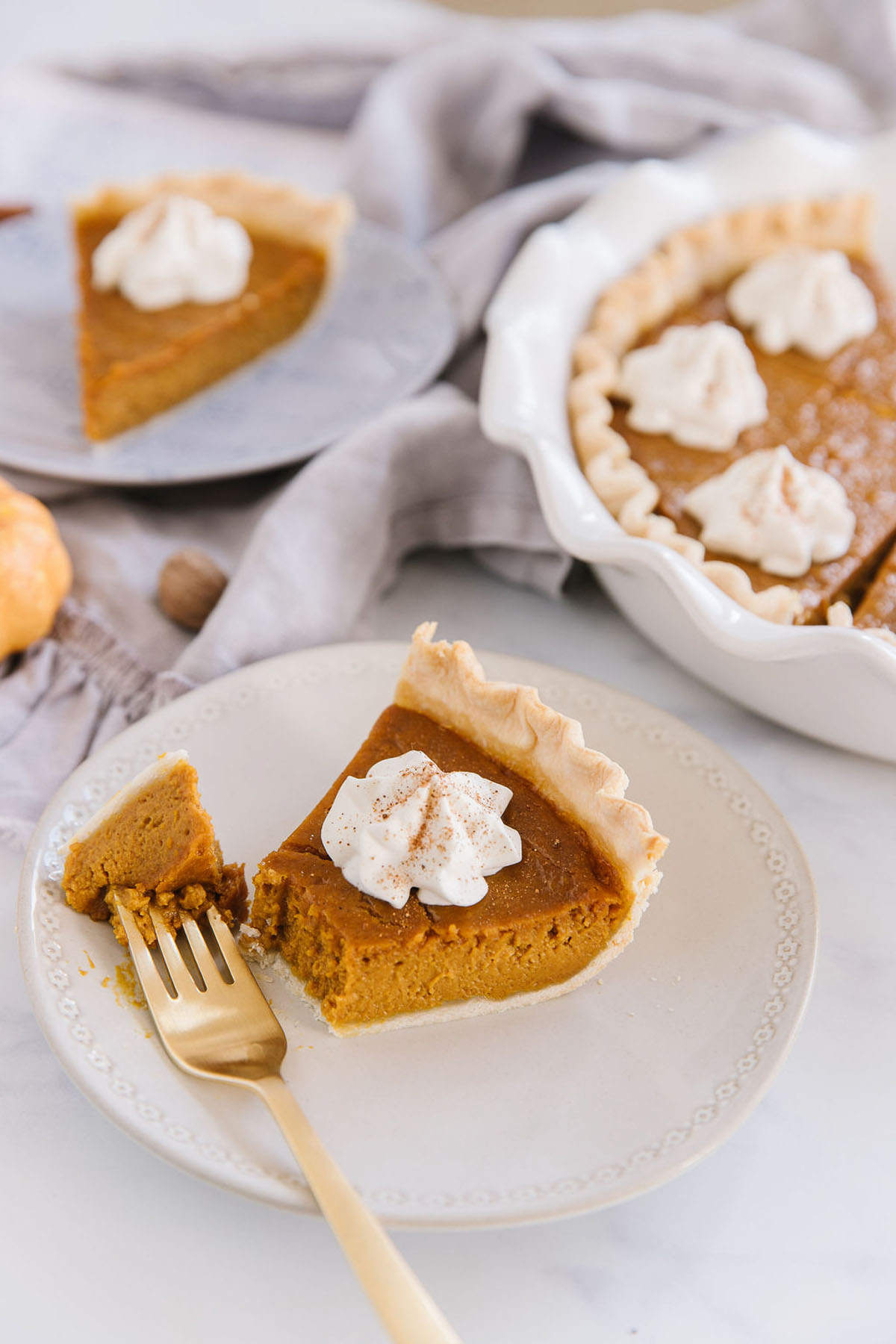 A white plate with fresh pumpkin pie slice with piece of it on a gold fork. Behind is a blue plate holding another pumpkin pie slice and a white pan of other pumpkin pie slices.