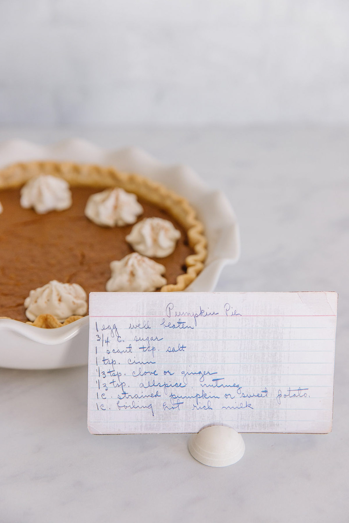 A white pan holding a fresh pumpkin pie with whipped cream dollops on it with a handwritten recipe card in front.