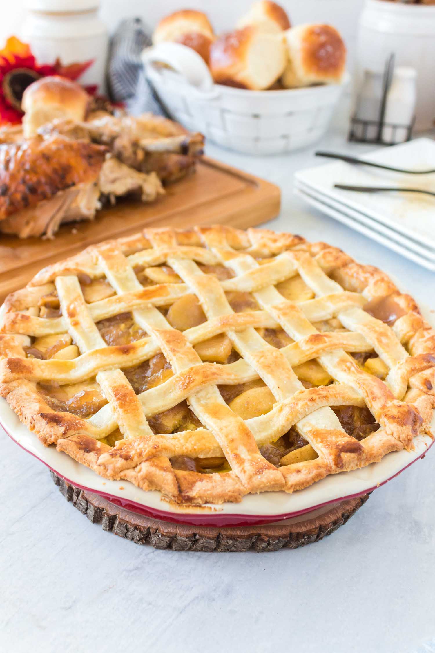 A whole homemade apple pie with turkey, rolls and plates in the background.