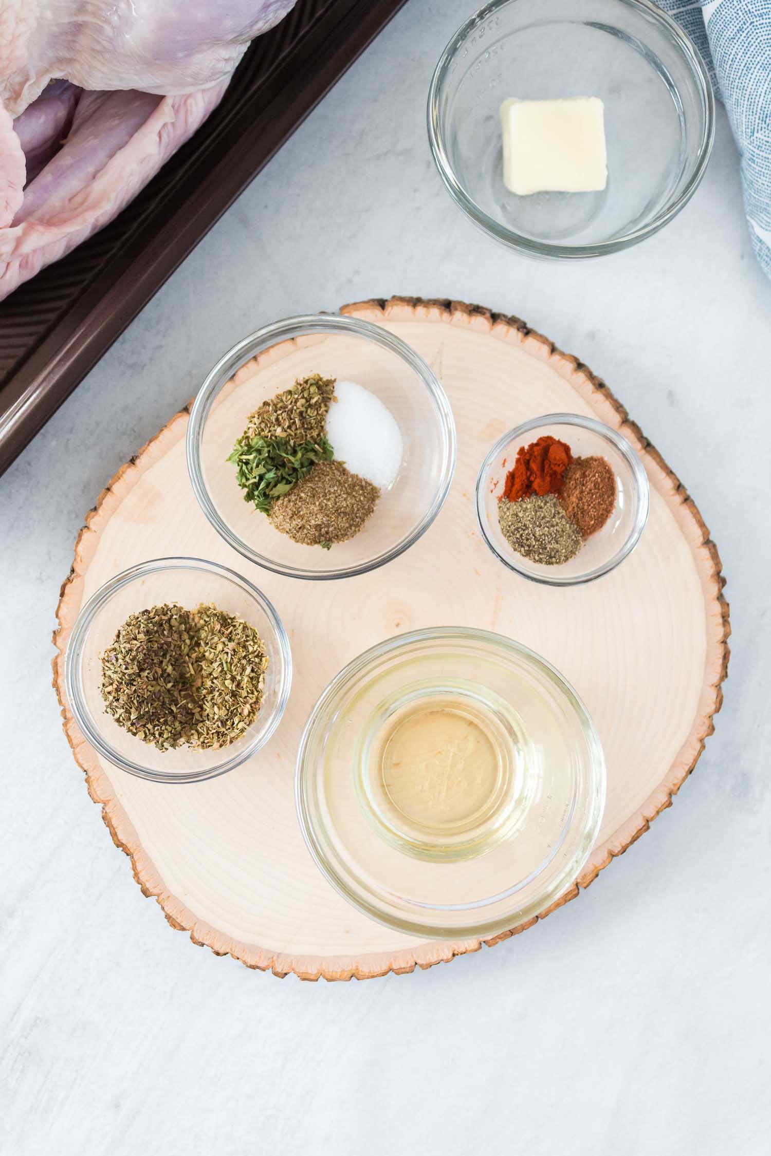 A wood board with four glass bowls filled with spices on it  and a glass bowl with a pat of butter on the side.