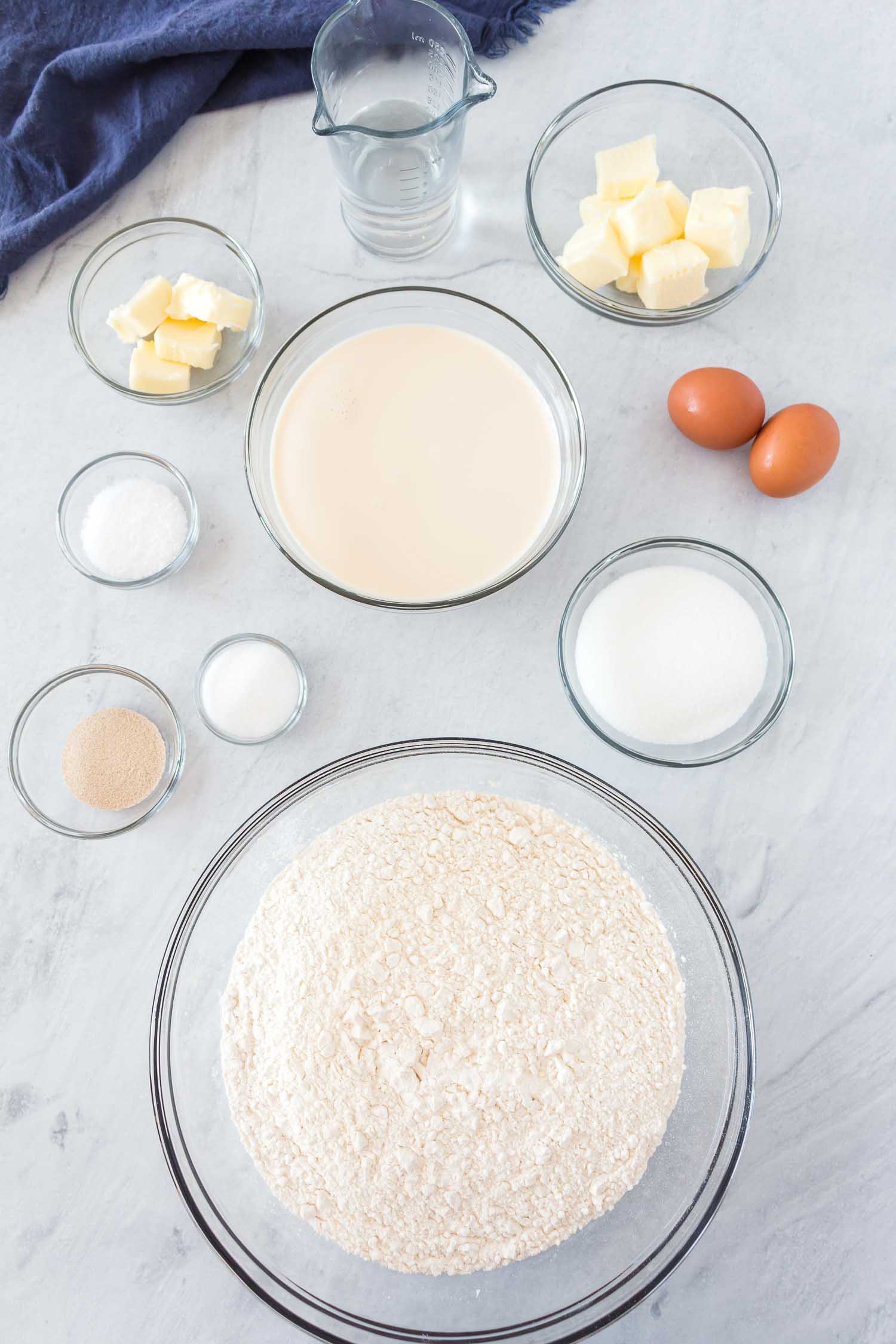 Glass bowls with ingredients: butter milk, flour, sugar, salt, yeast, water and 2 whole eggs on the side.
