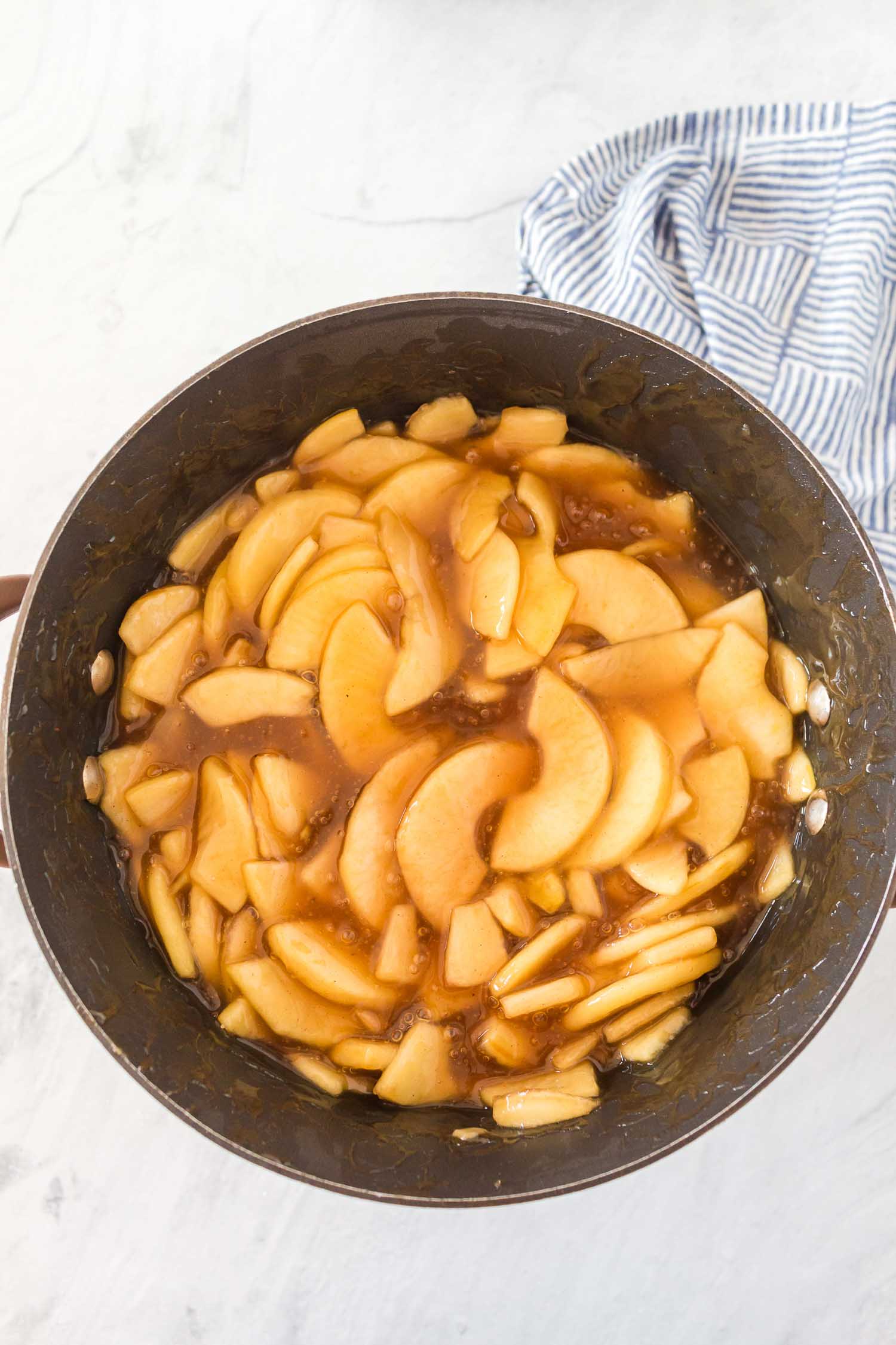 A crock pot full of cooked, sliced apples and spices with a blue and white striped towel in the corner.
