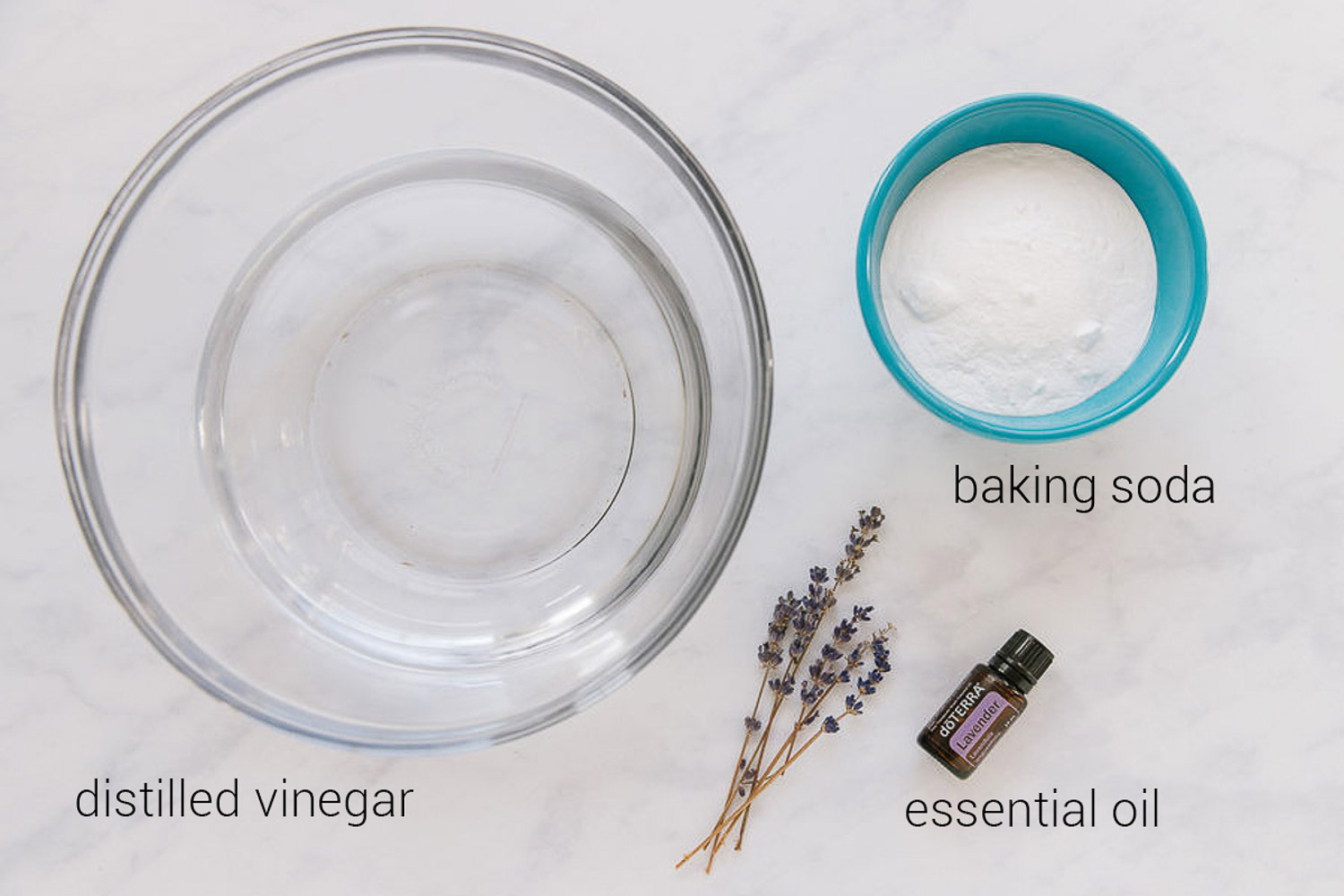 A glass bowl with vinegar in it,  a blue bowl with baking soda in it and a bottle of lavender Doterra essential oil and a piece of lavender by it.