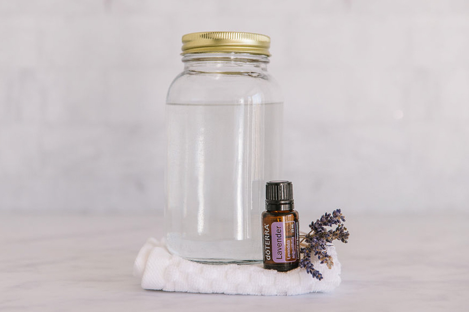 A clear mason jar filled with vinegar on top of a white towel with a bottle of lavender Doterra essential oil and a piece of lavender.