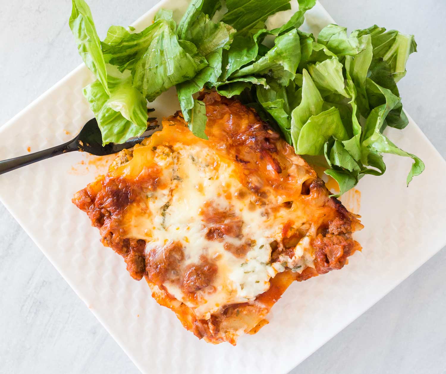 A white plate of a lasagna serving with lettuce on the side and a black fork.