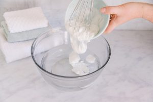 Person using a whisk to move conditioner from a small bowl to a large glass bowl of water.