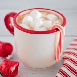 Candy cane hanging on the side of a mug of hot chocolate topped with marshmallows.