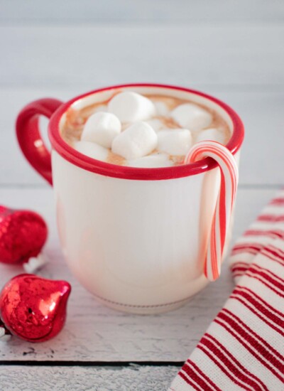 Candy cane hanging on the side of a mug of hot chocolate topped with marshmallows.