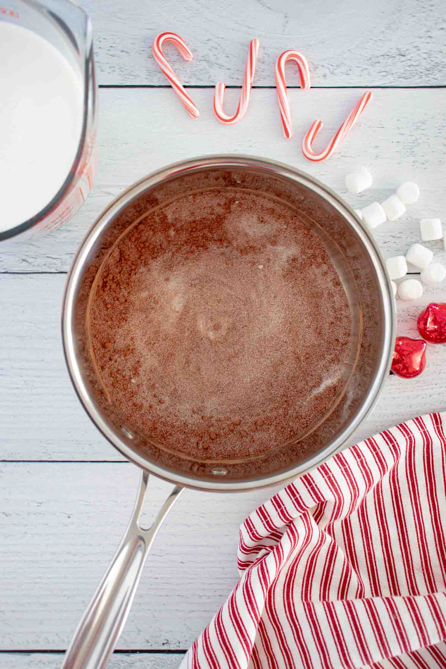 A metal pan with hot chocolate in it with a measuring cup of milk, candy canes, marshmallows, ornaments and a red striped towel next to it.