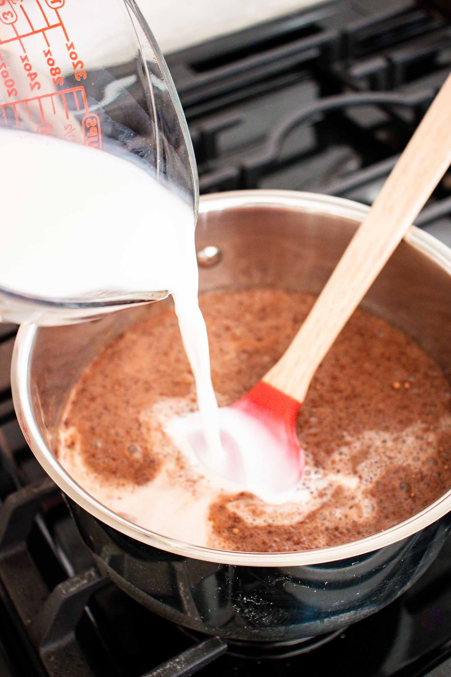 A metal pan with hot chocolate in it with milk pouring into the pan. there is a red spatula in the pan.