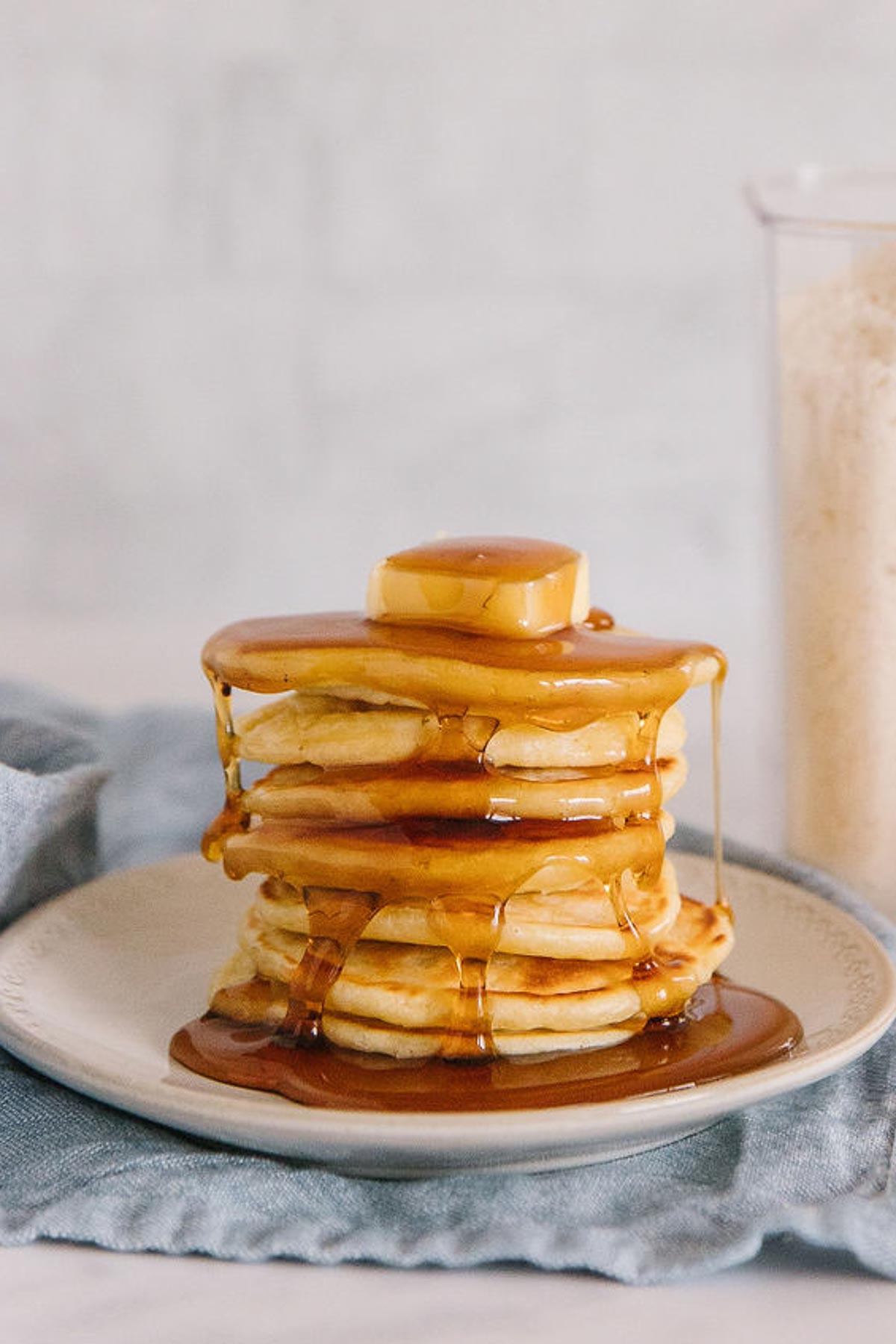 A stack of pancakes with a pat of butter on top and syrup all over it on a plate with a plastic container of bisquick mix on the side.