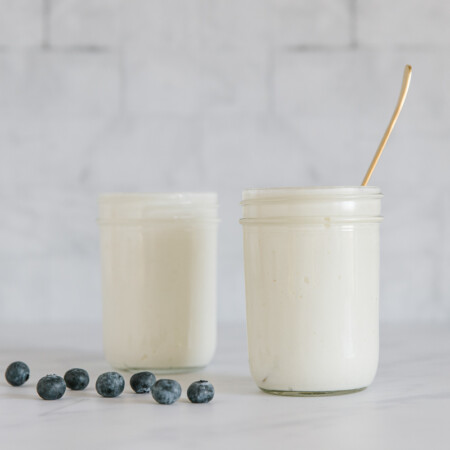 Two jars of Greek yogurt on a counter with blueberries.