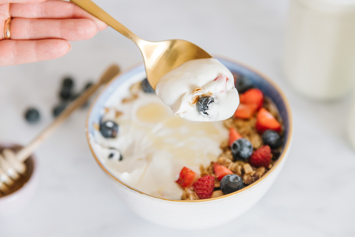 A white bowl of yogurt with fruit and granola on it with a gold spoon with yogurt and a blueberry on it. There is a honey stick and blueberries on the counter.