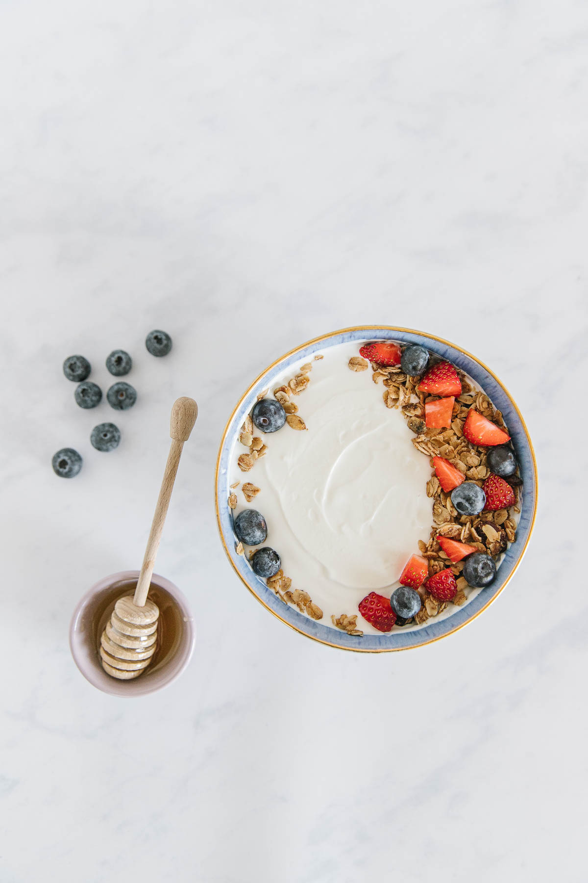 A blue-rimmed bowl of yogurt with fruit and granola on it with a honey stick in a bowl of honey and scattered blueberries on the counter next to it.