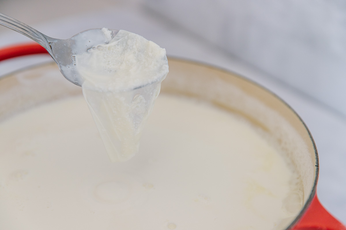 An orange Dutch oven filled with a white mixture with a metal spoon bringing up the skin of the milk.