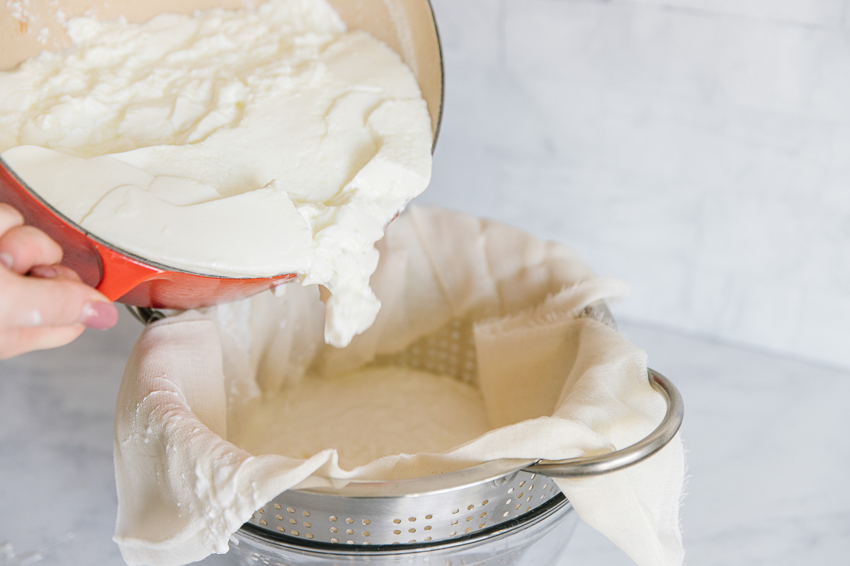 An orange Dutch oven is pouring white liquid into a metal colander with cheesecloth in it.