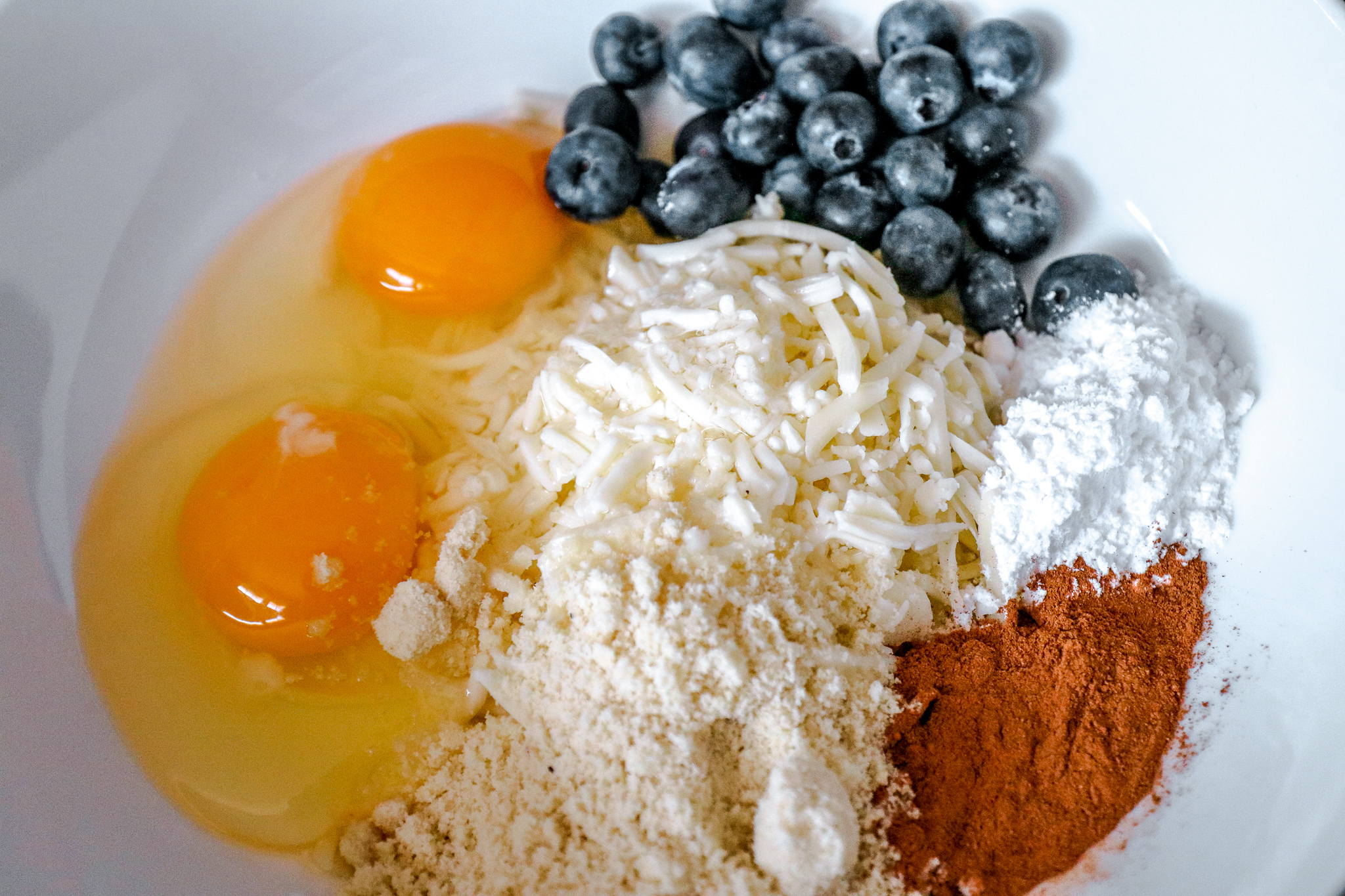 A white bowl with ingredients for chaffles - eggs, almond flour, blueberries, cinnamon, cheese and swerve.