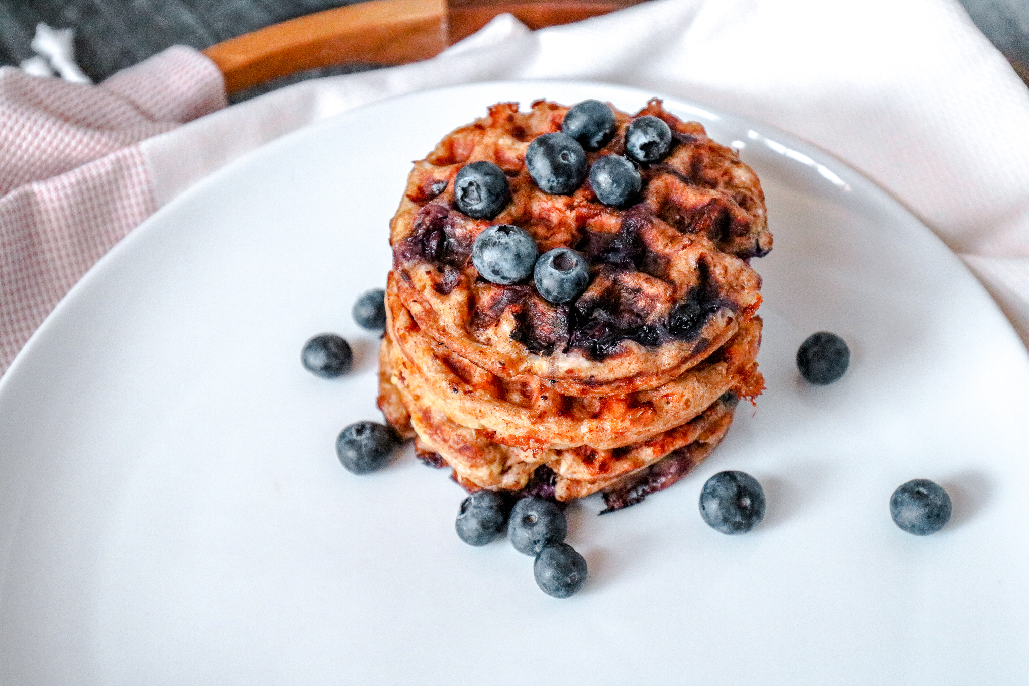 A white plate with four mini chaffles stacked on top of it with blueberries on top and sprinkled on the plate on top of a towel underneath.