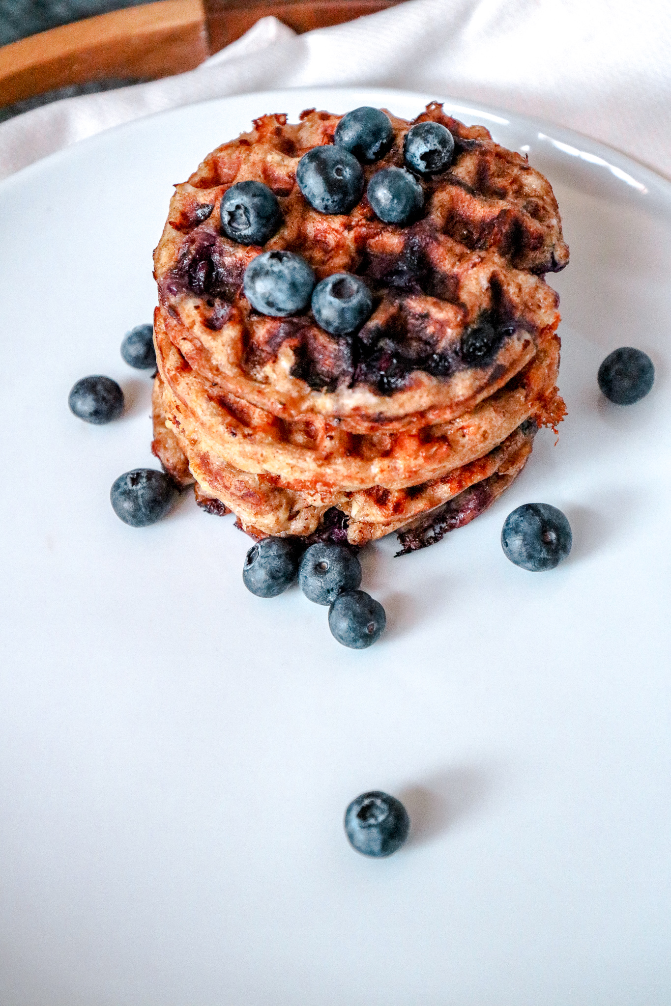 A white plate with four mini chaffles stacked on top of it with blueberries on top and sprinkled on the plate.