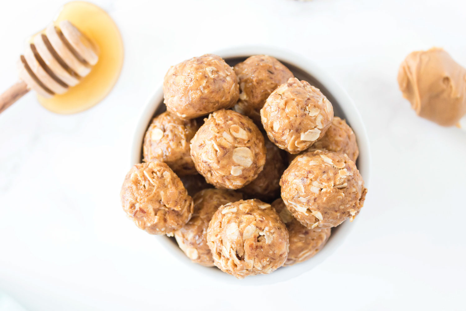 A bowl of energy balls with a dollop of honey and want and a spoon of peanut butter on the side. 