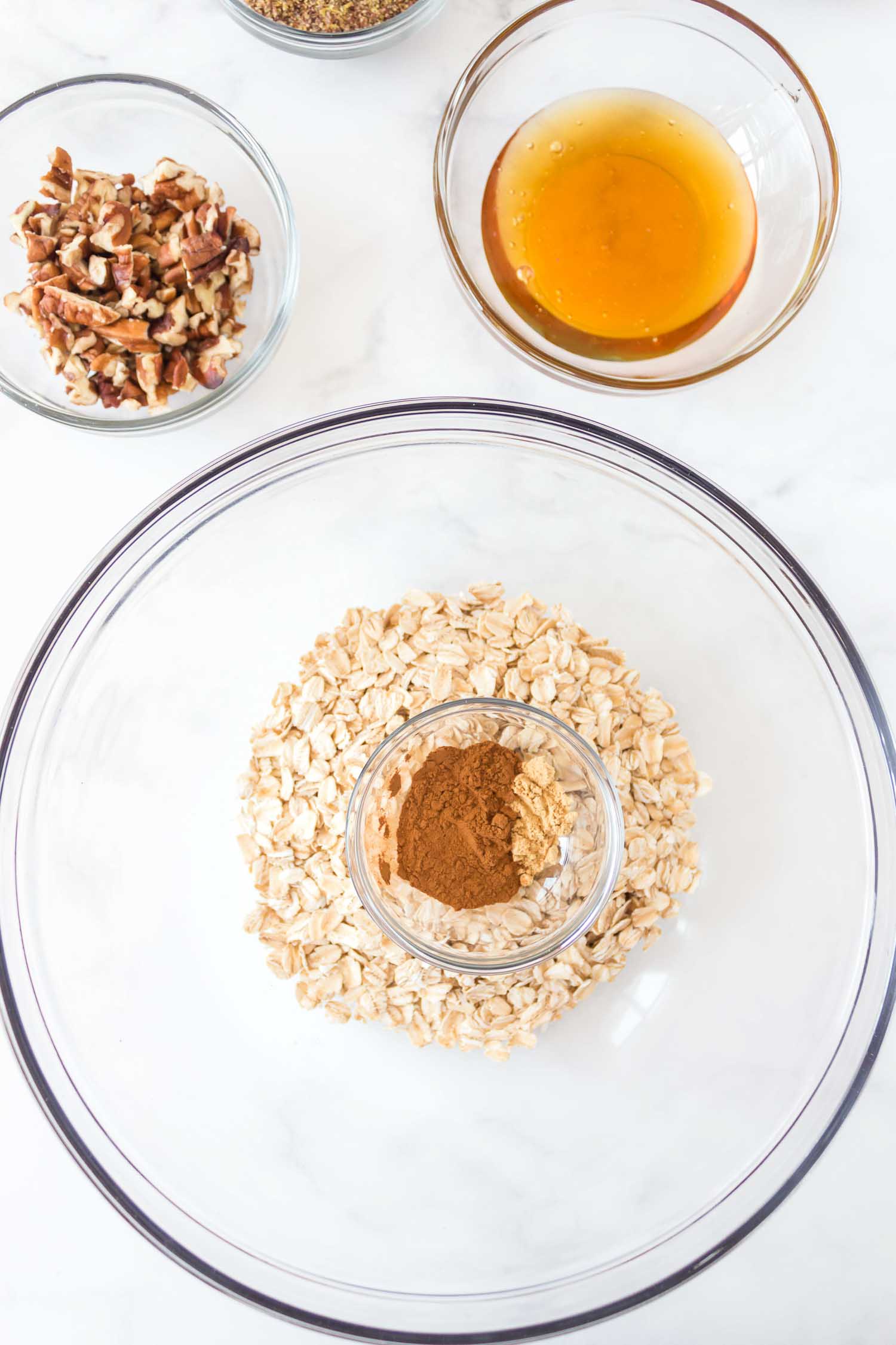 A large glass bowl of oatmeal with a smaller glass bowl of spices on top of it with smaller glass bowls of honey and pecans behind it.
