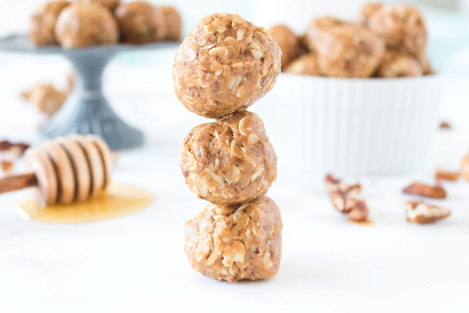 Three energy bites on top of each other with a pile of honey and wand and pecans on the ground. There is a white bowl and a pedestal holding protein balls on it.