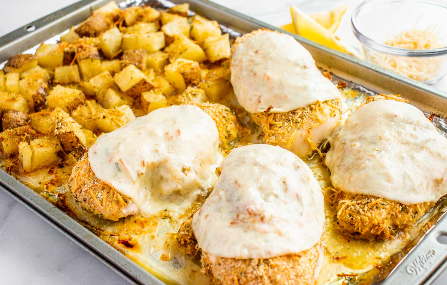 A metal pan with cooked potatoes and 4 chicken breasts with melted cheese on top of it.