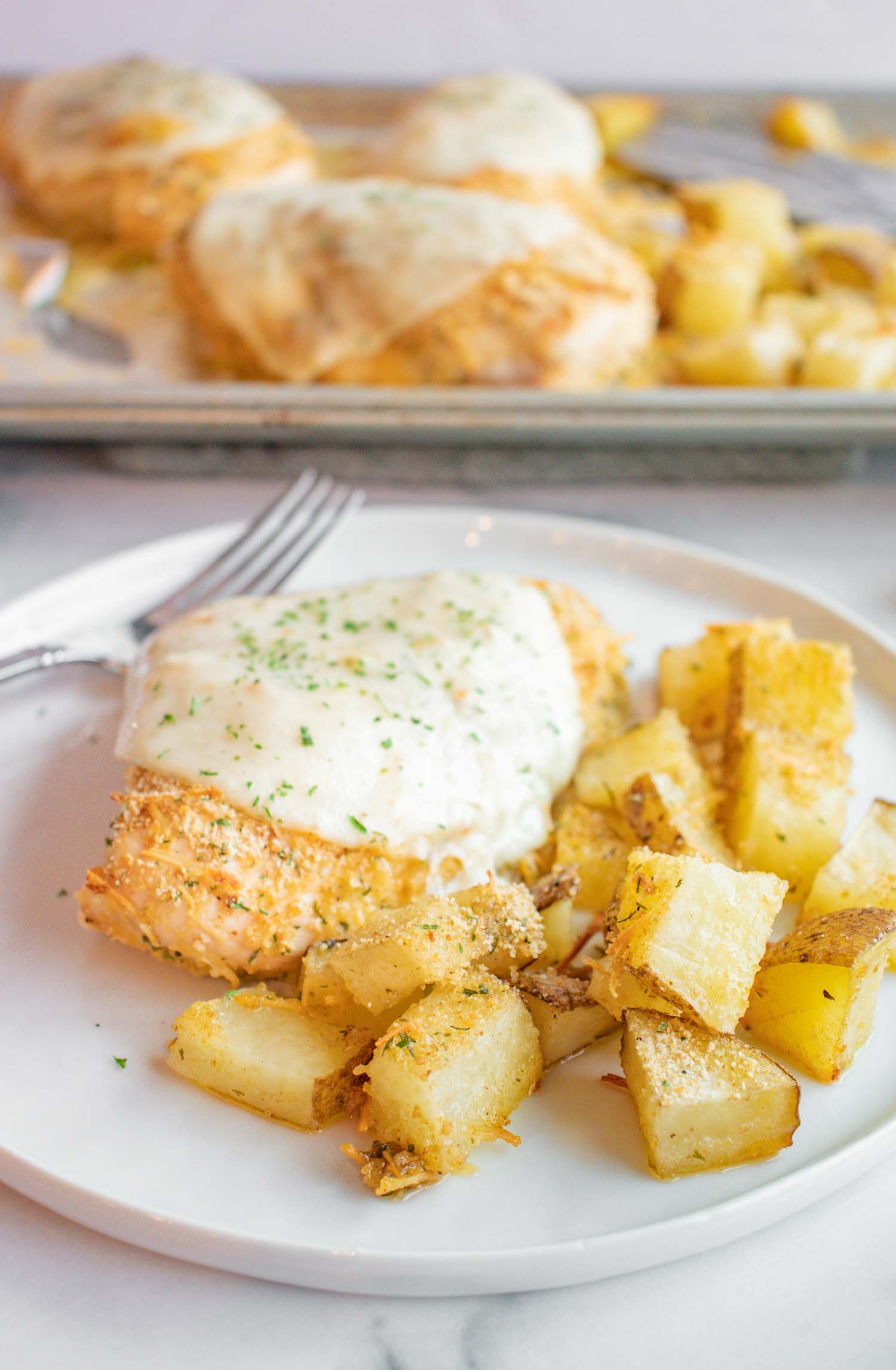 A white plate with a piece of chicken with cheese on it with cubed potatoes on the side with a metal fork and behind is a metal pan with more chicken and potatoes on it.