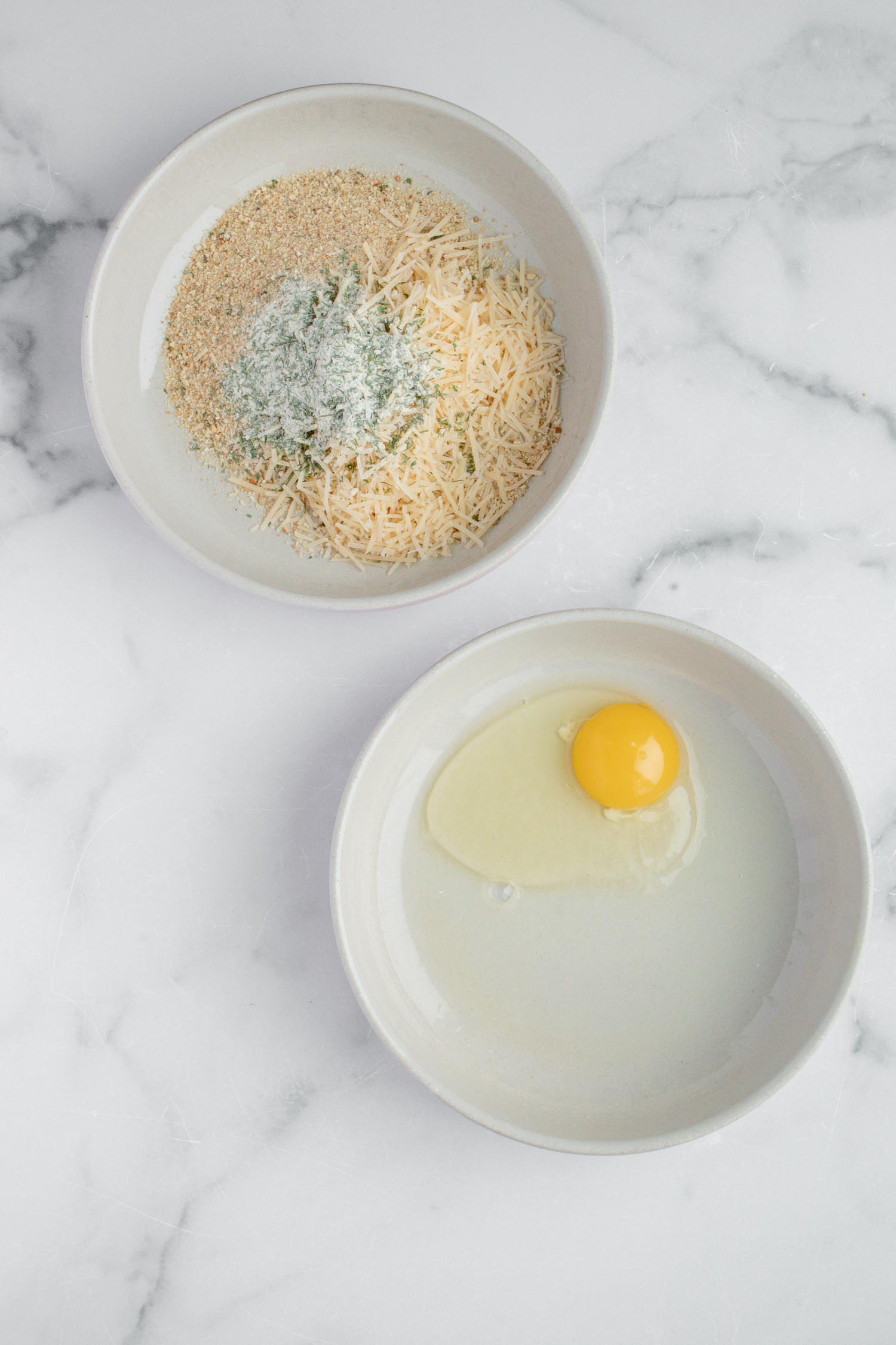Two white bowls with an raw cracked egg in one and in the other is spices and cheese.