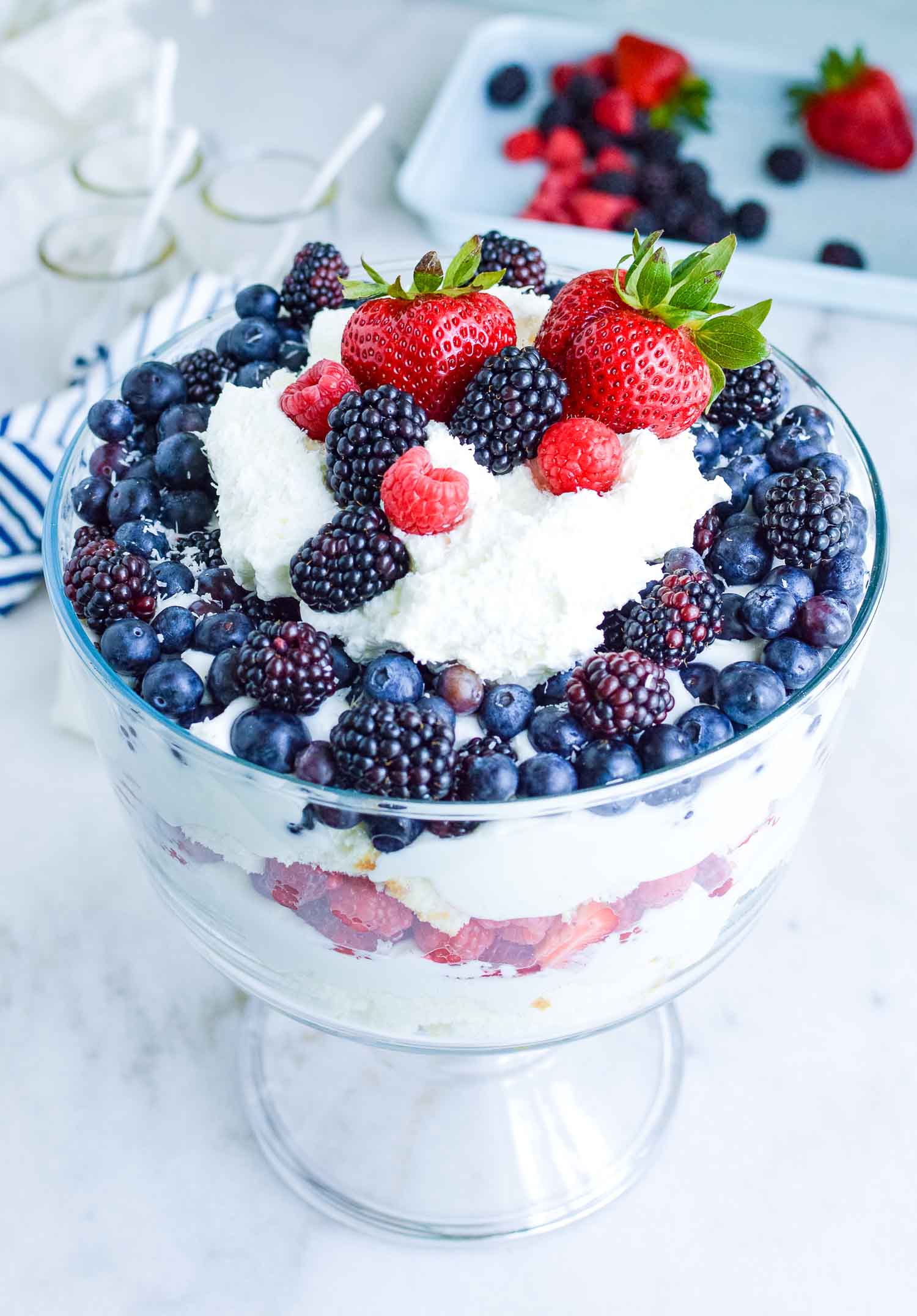 A clear glass dish filled with cake, berries, and cream with a blue and white towel behind it with a plate of extra berries and three empty glass cups with white spoons in them.
