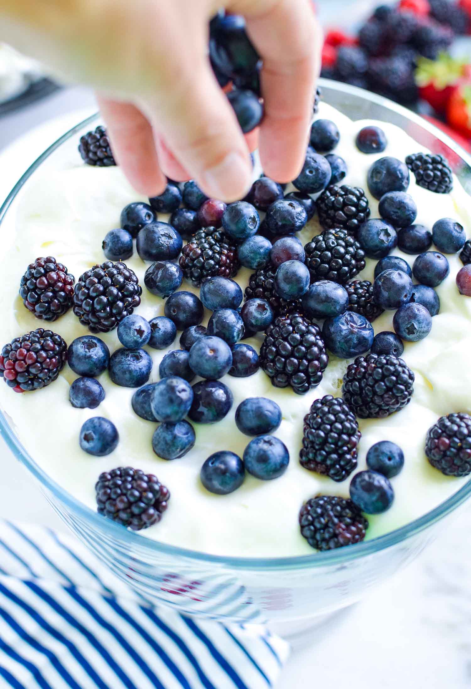 A clear dish with a topping of cream and blue assorted berries being added by a hand on top.