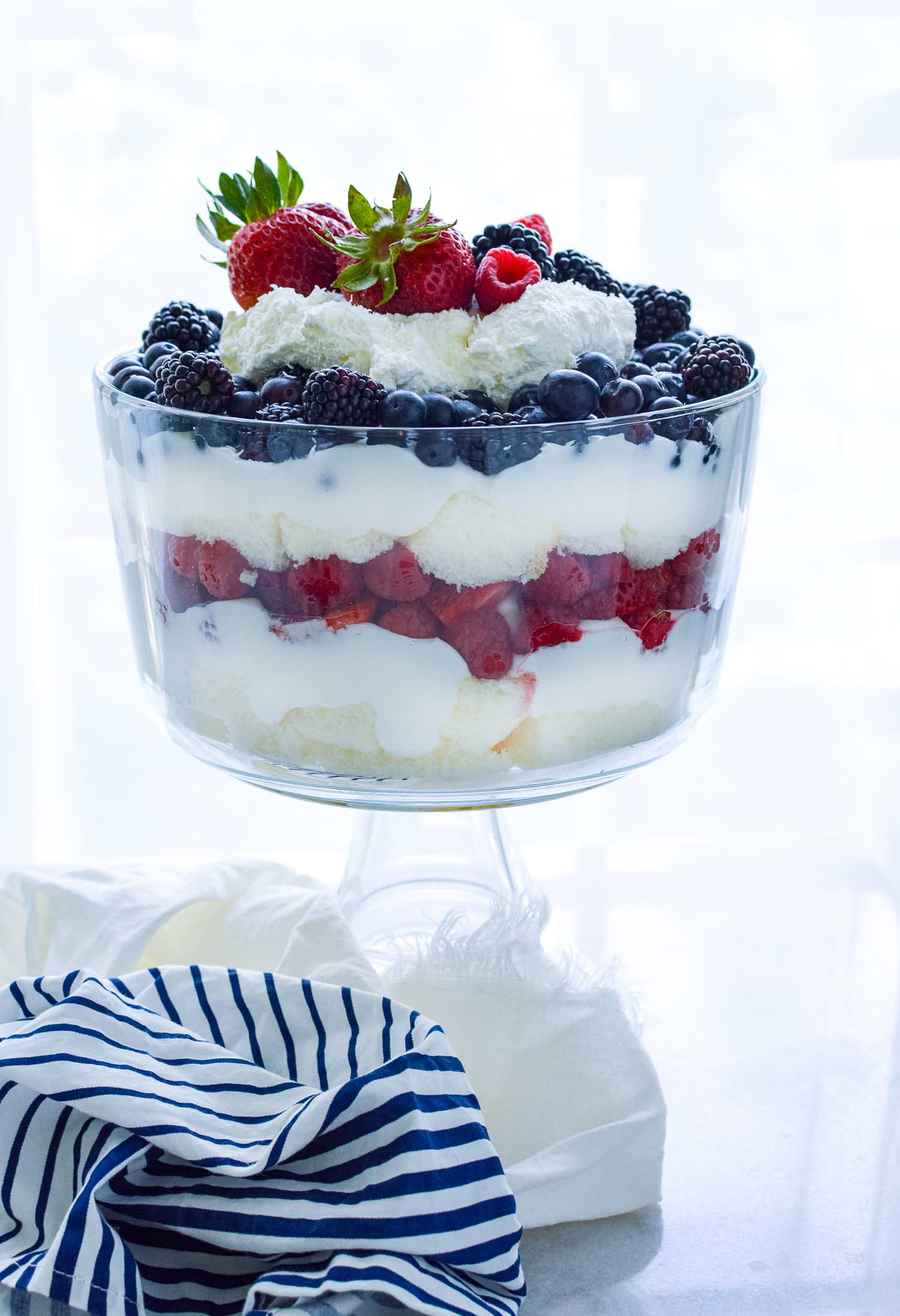 A clear glass trifle dish filled with cake, berries, and cream with a blue and white towel around the bottom.