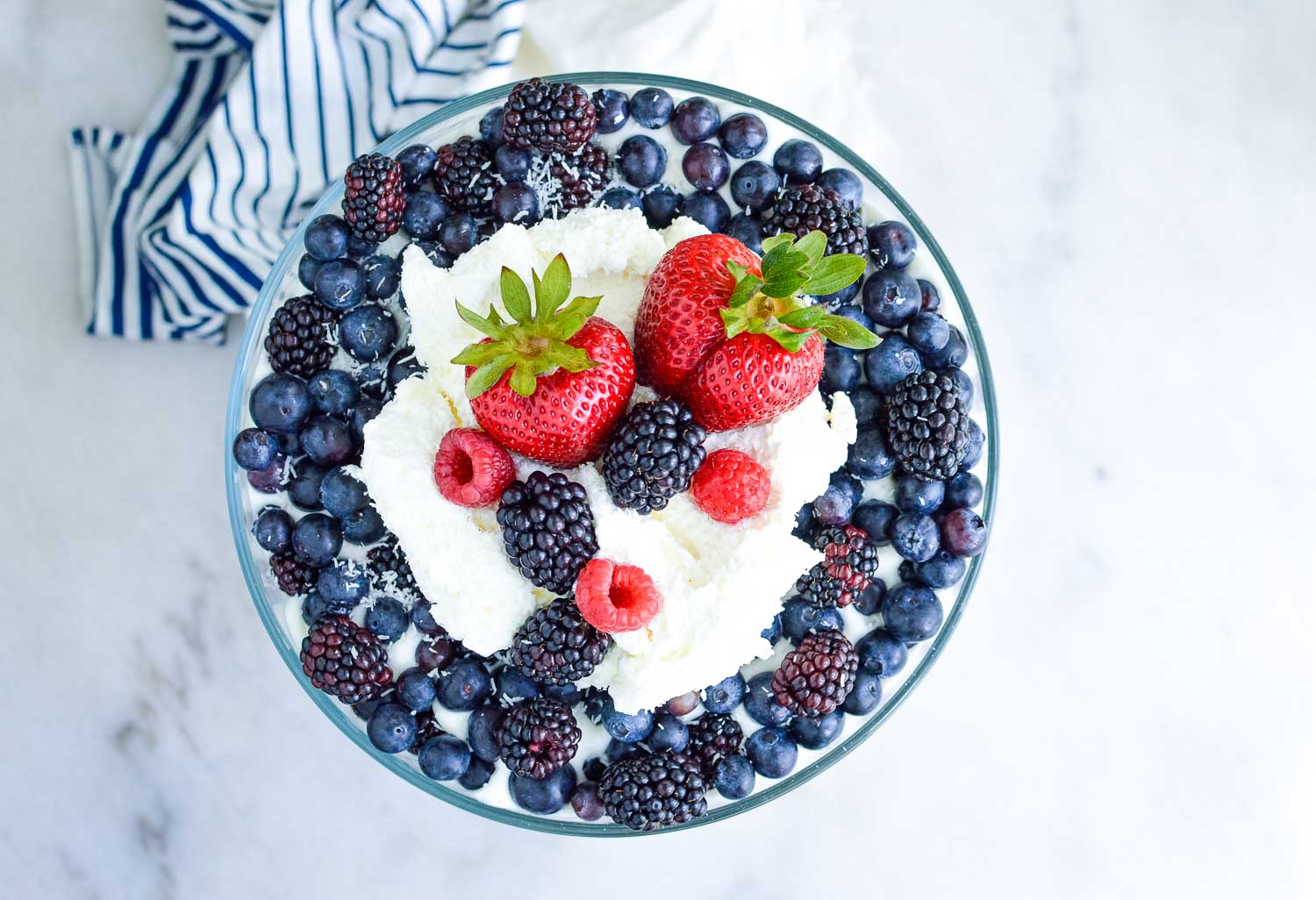 A top view of a clear glass dish filled with cake, berries, and cream with a blue and white towel around the bottom.