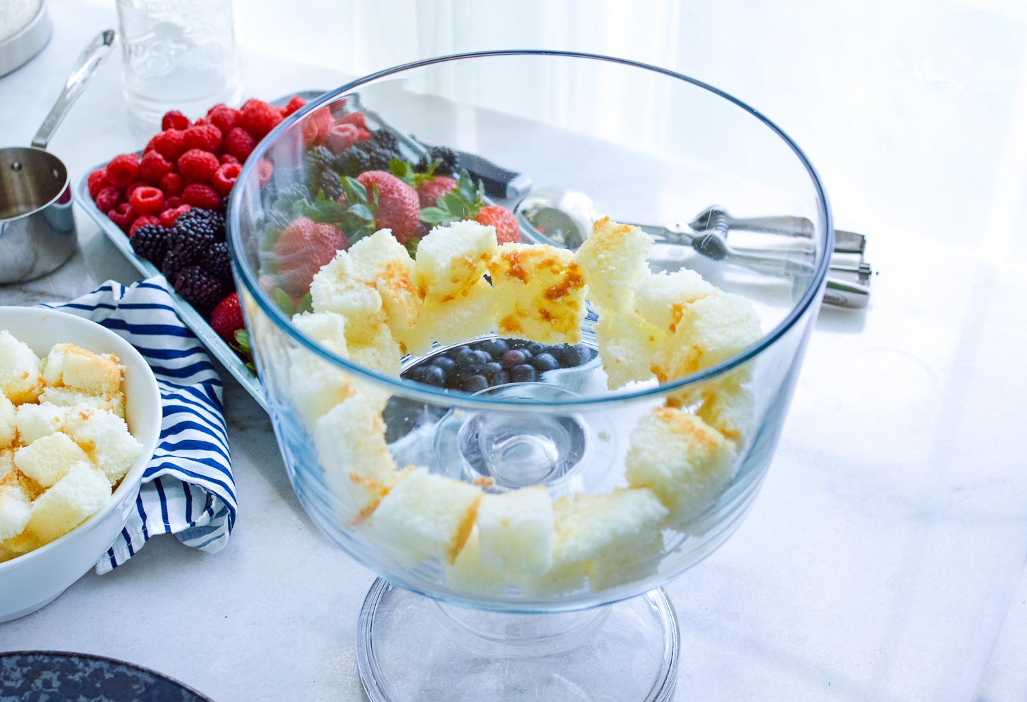 A mostly empty clear glass trifle dish with cut cake around the sides with a bowl of more cake and a plate filled with berries next to it with a metal measuring cup next to it.