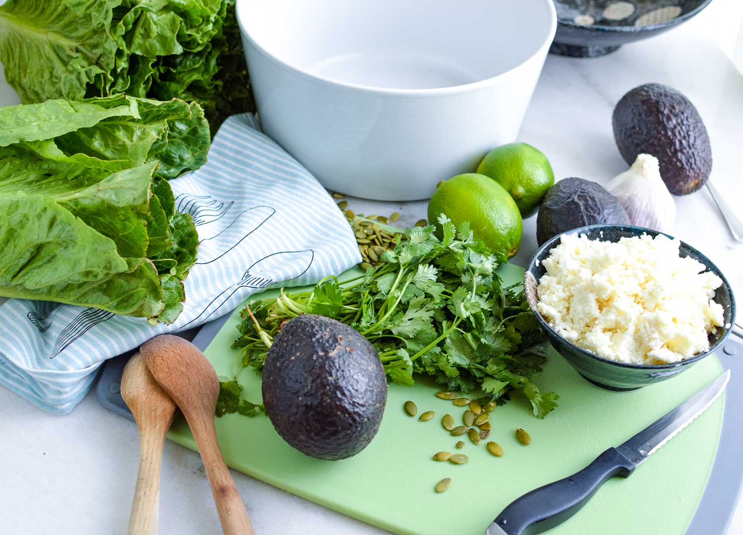 An empty white bowl with ingredients on the side: romaine lettuce, limes, cilantro, avocados, cheese on a cutting board and a knife.