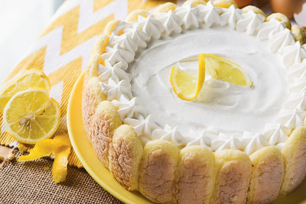 A whole cake on a yellow plate with a slice of lemon on top and also on the sides with cut lemons on a yellow and white chevron towel.