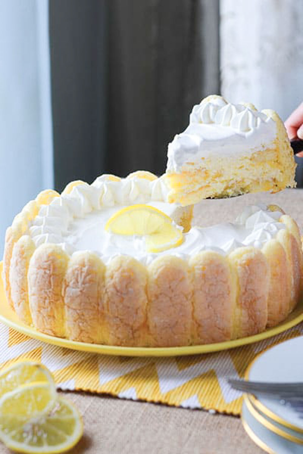 A whole cake on a yellow plate with a slice of cake being lifted by a hand on a black spatula above it.