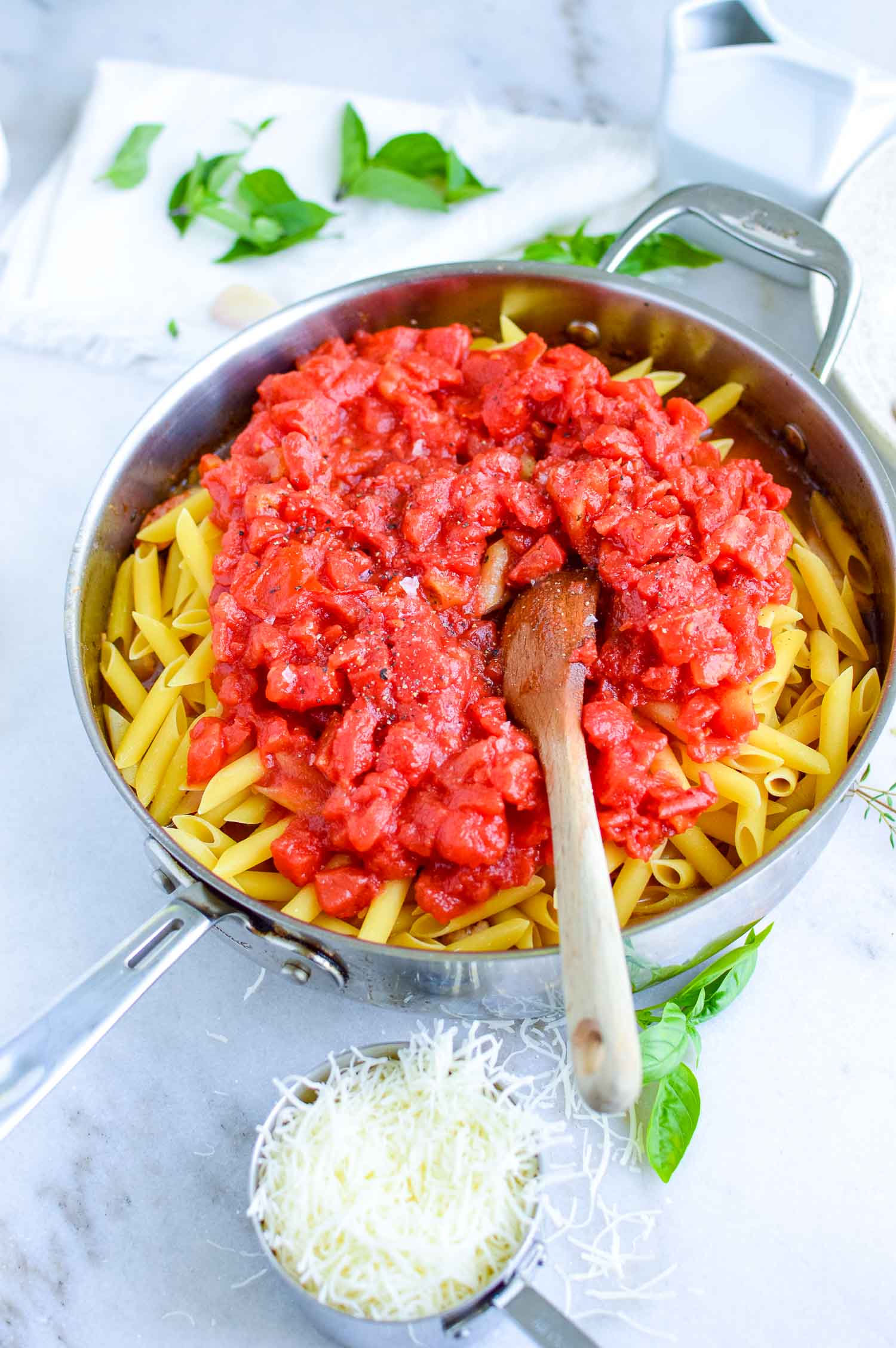 A silver pan of uncooked penne pasta with cut tomatoes and spices along with a wooden spoon inside of it and fresh herbs around it.