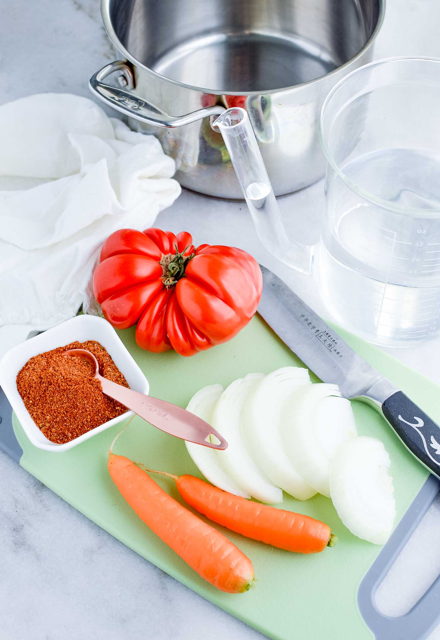 A silver saucepan, tomato, seasonings, water, onion, carrot and a plastic container of water with a green cutting board and knife and a white towel.