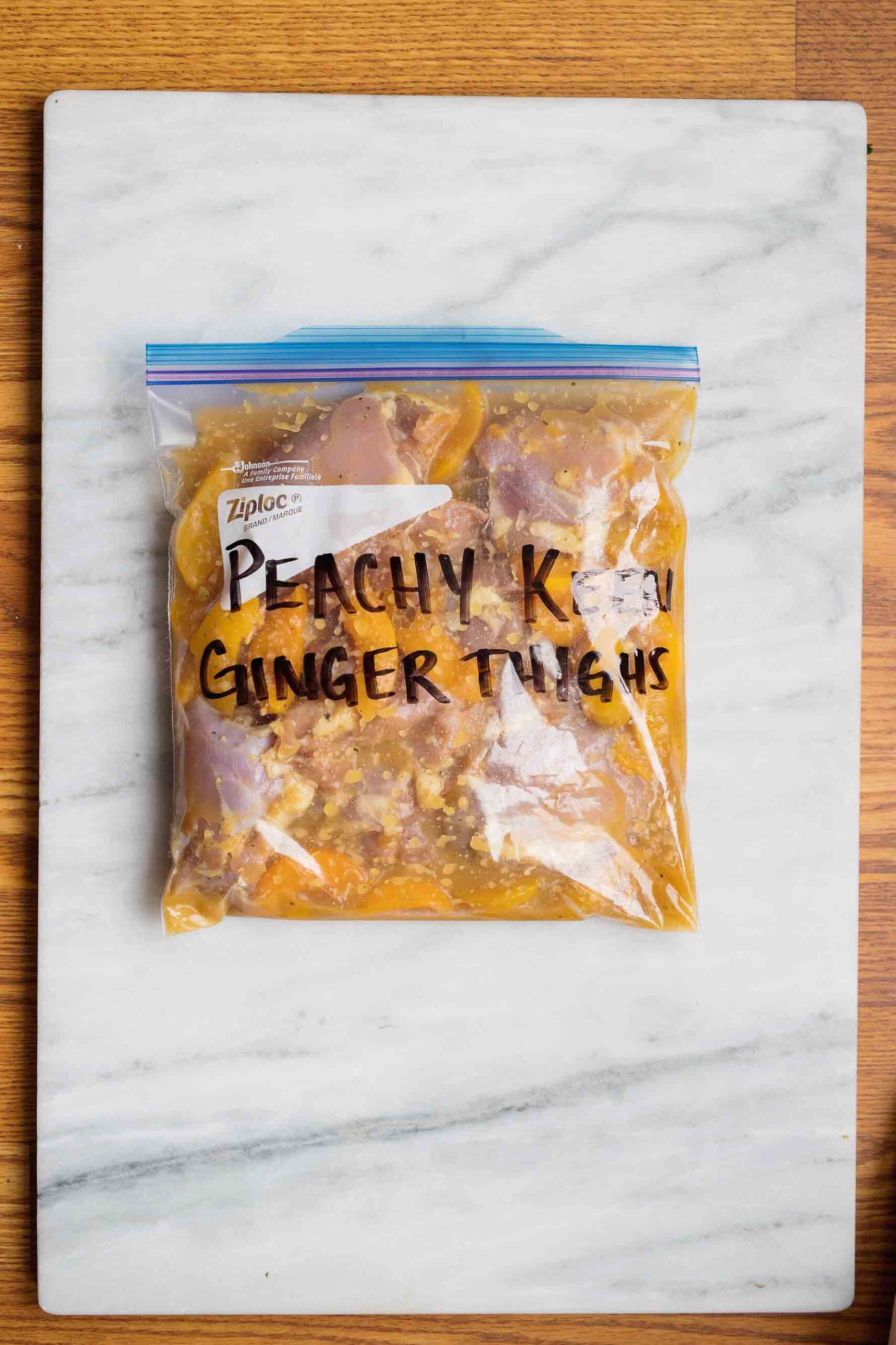 A resealable bag with raw chicken and sauce in it with the words peachy keen ginger thighs written in black on it on a whit marble cutting board.