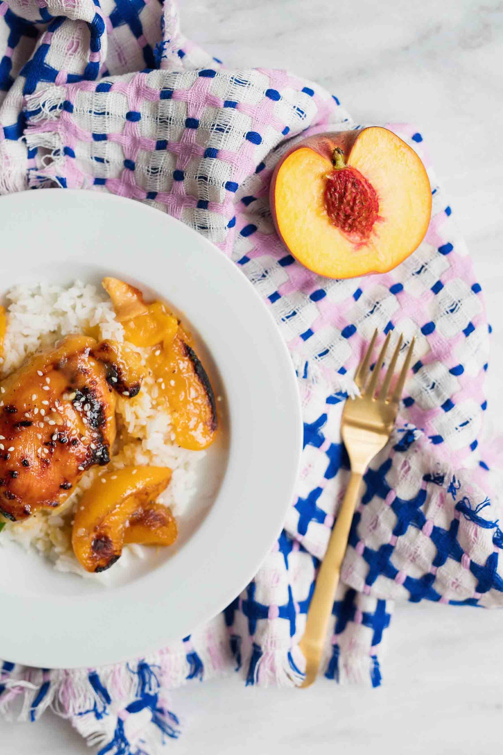 A white bowl with white rice and peaches with cooked thighs on it. There is a blue and pink cloth with a gold fork and half of a peach on it.