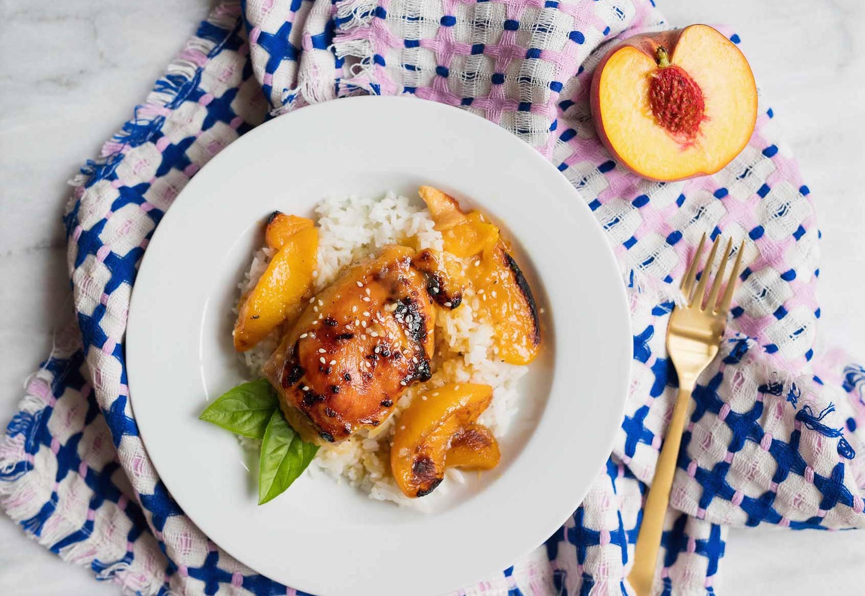 A white plate with rice and peaches with chicken on it. There is a blue and pink cloth with a gold fork and half of a peach on it with a white marble background.