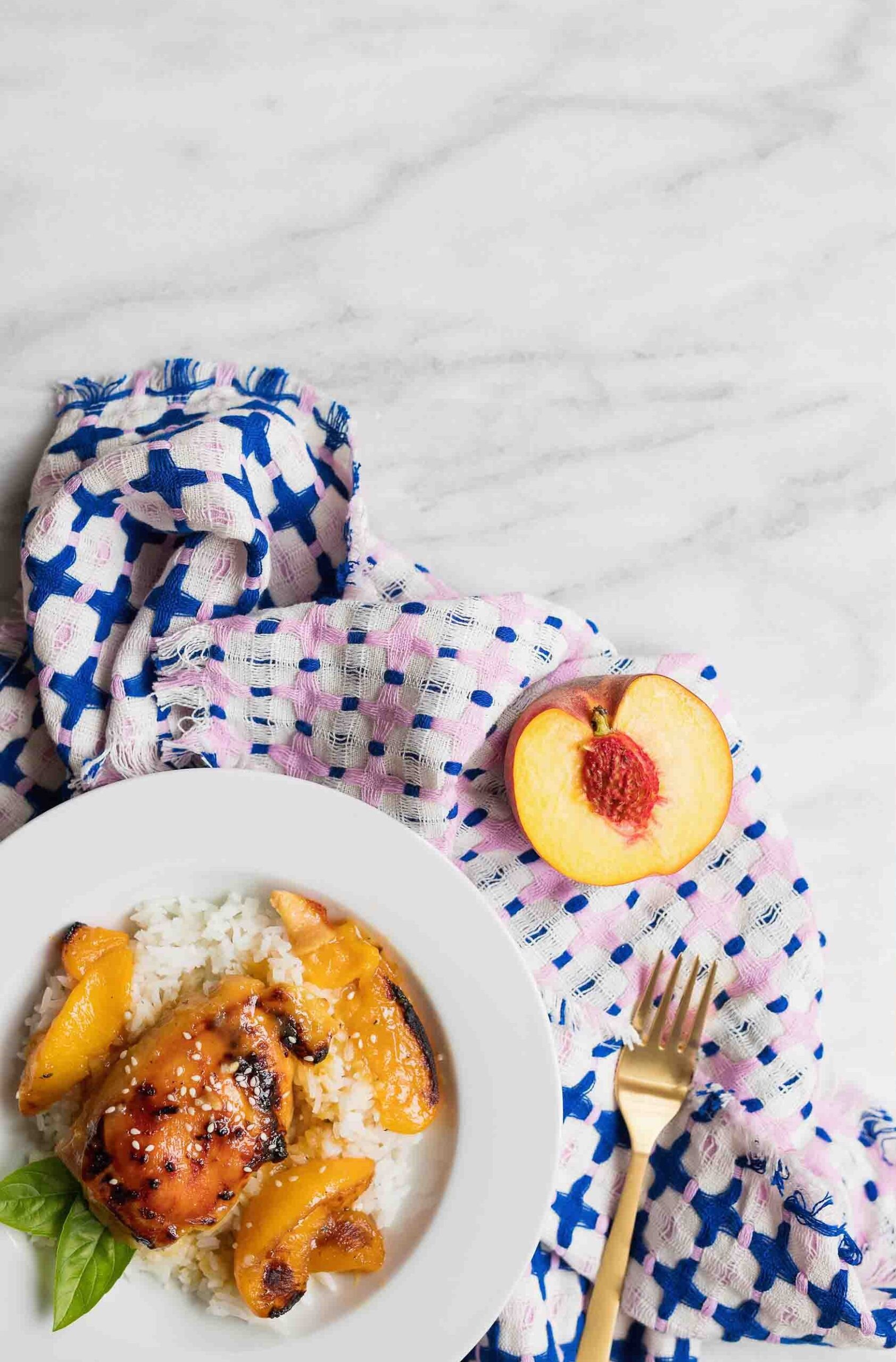 A white plate with rice and peaches with chicken thighs on it. There is a blue and pink cloth with a gold fork and half of a peach on it on a white marble counter.