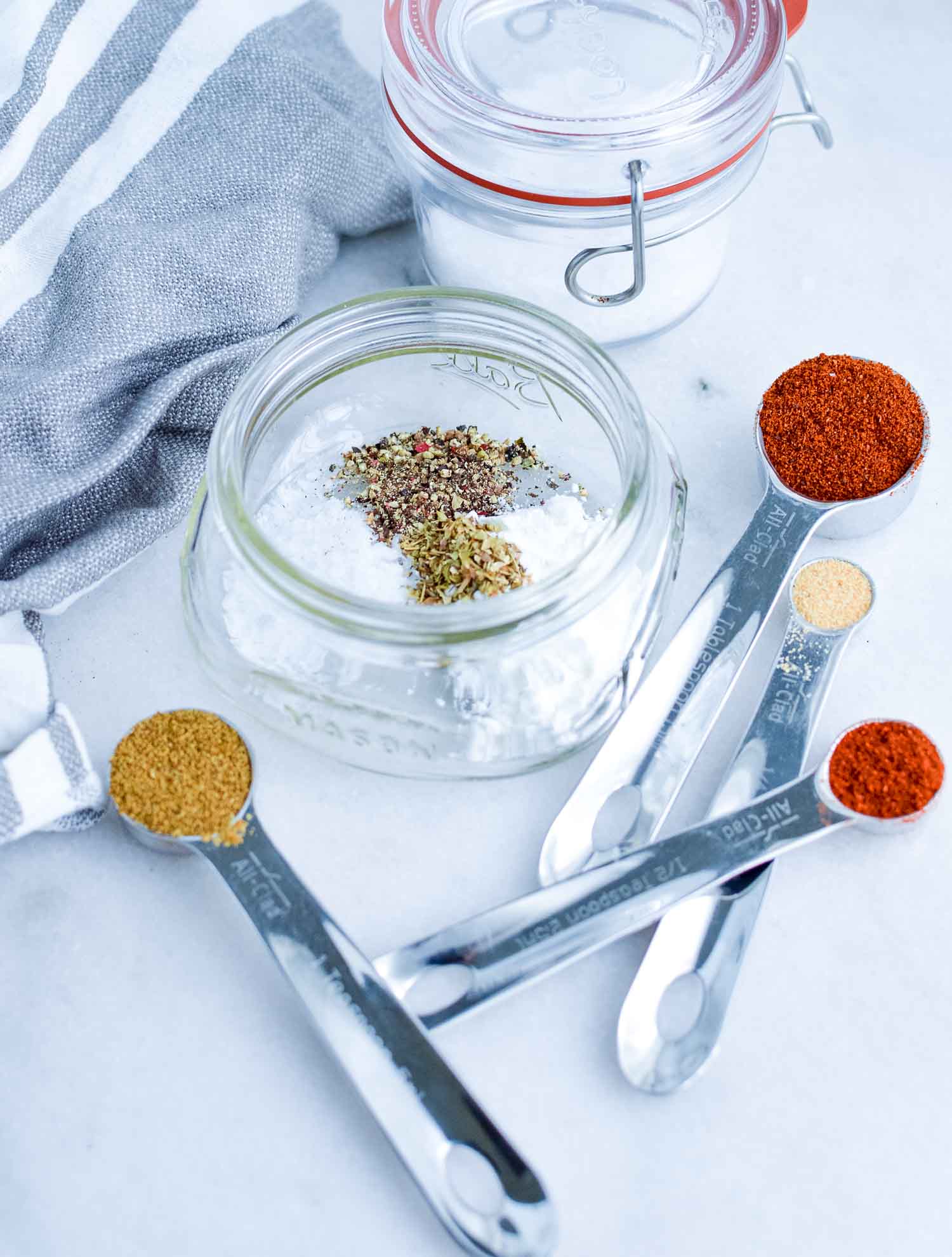 A glass jar with spices in it with 4 silver measuring spoons with spices filling them.