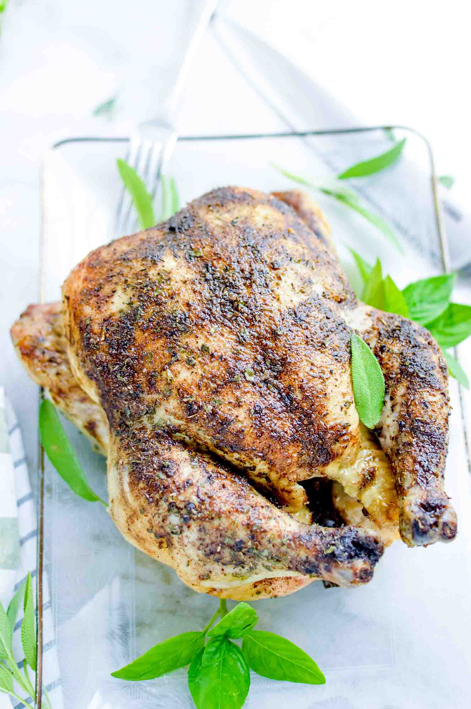 A whole roasted chicken on a clear tray with a metal knife and green herbs around it.