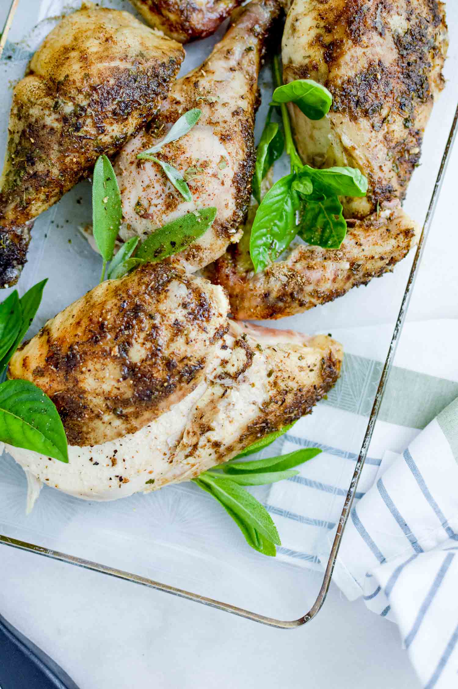 A clear tray with a cut seasoned chicken and green herbs all on and around it with a blue and white striped towel next it.